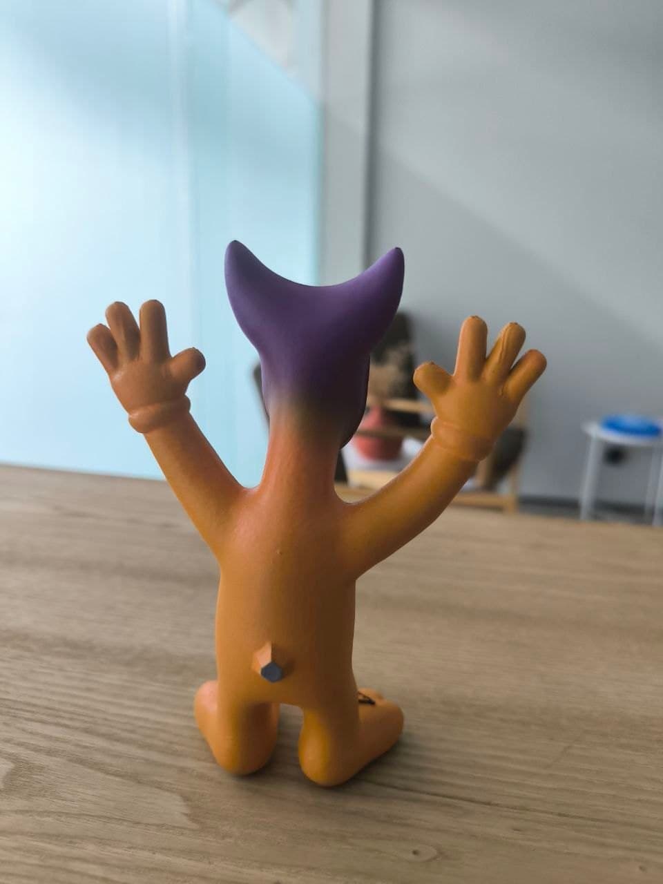 Finished painted Apophis prototype, back view: orange body, raised hands, purple horns rising from behind the head, on a wooden tabletop.