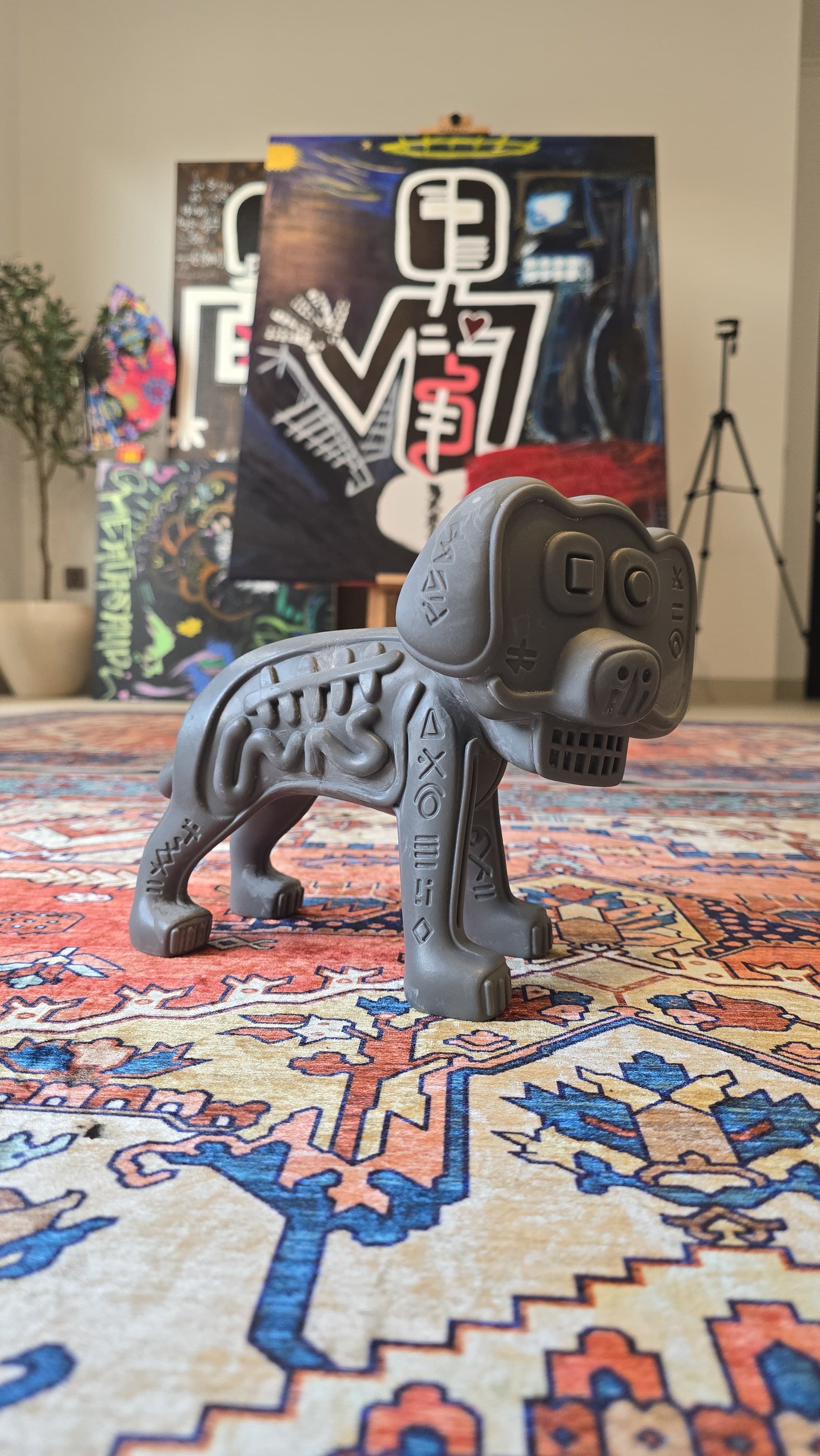 Raw 3D-printed Marley prototype in gray resin standing on a Persian carpet — a skeletal dog with long drooping ears, ribs and an intestinal coil sculpted into the flank, and glyph rows cut into the legs, with a dark glyph-marked canvas on an easel behind.