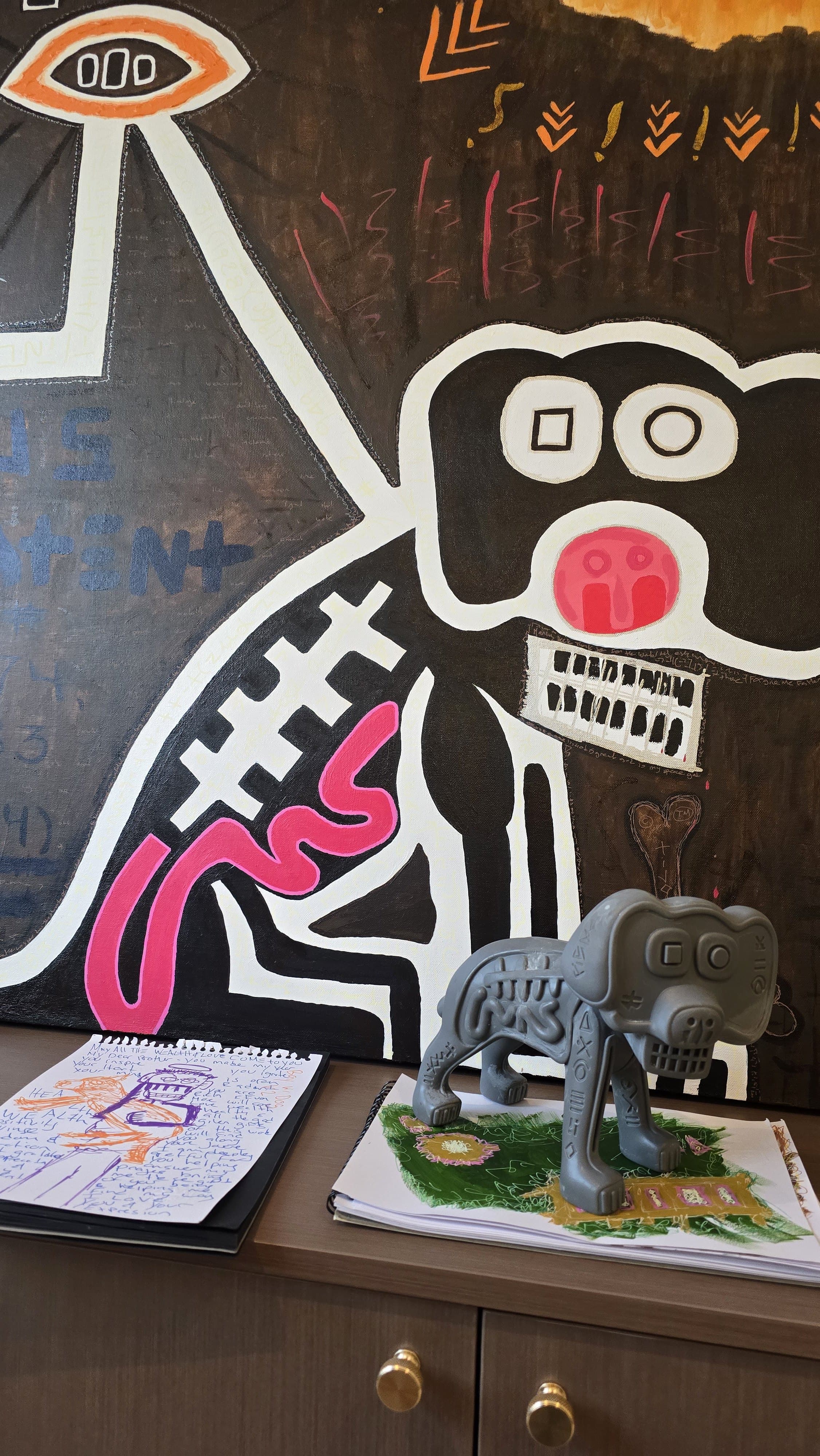 The raw gray Marley print standing on a sideboard directly beneath its source painting Boy & Dog V2 — the painted black dog with mismatched square and circle eyes, pink nose, white tooth-grid, and pink intestinal coil rising directly above the sculpt of the same character.