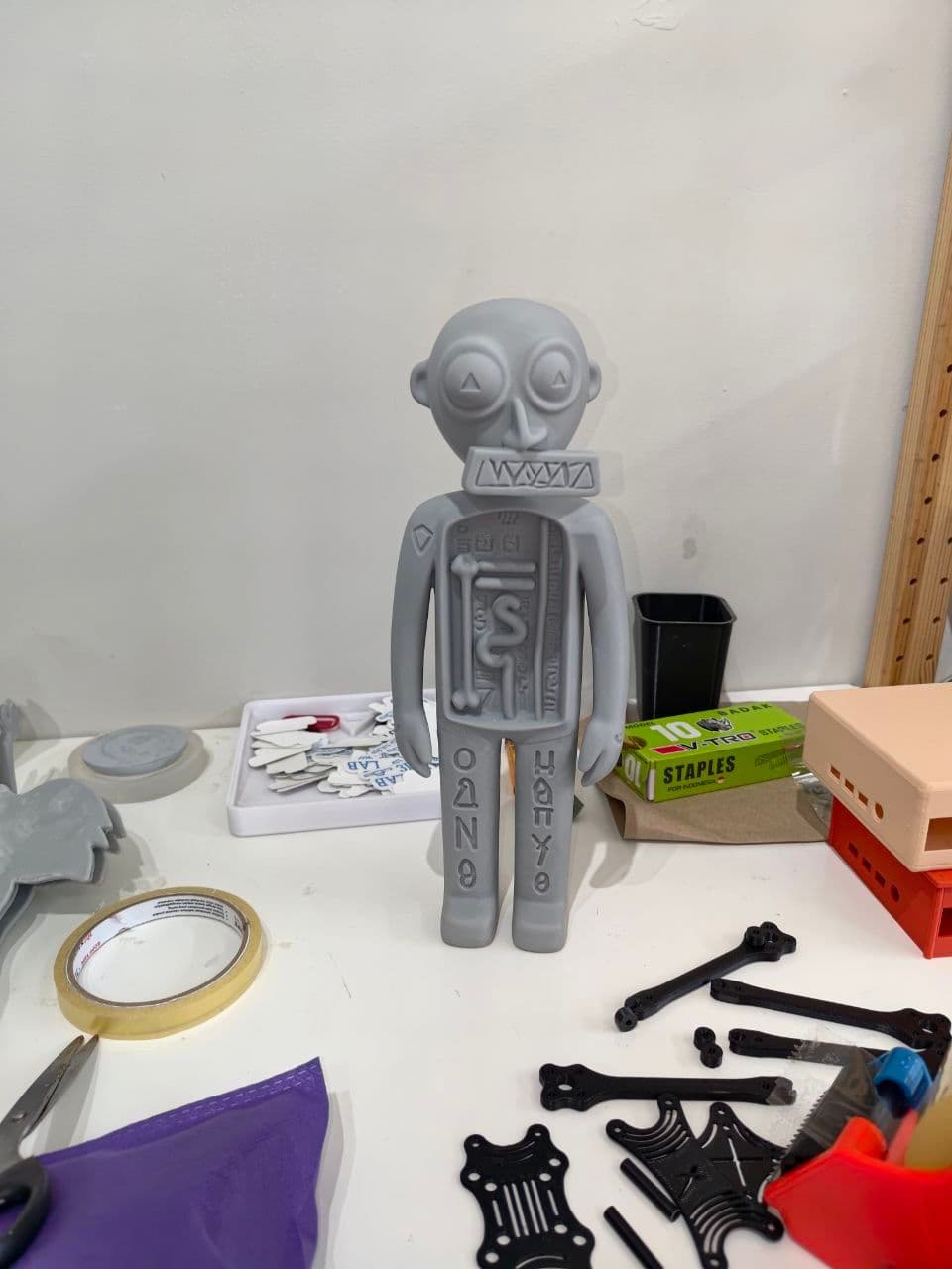 Raw 3D-printed prototype of a Three Angels figure standing on a white studio table: gray resin humanoid with round eyes holding triangle pupils, an elongated nose, a wide zigzag-patterned collar, and an open torso showing bone, serpent, and heart markings with rows of small glyphs running down both legs.