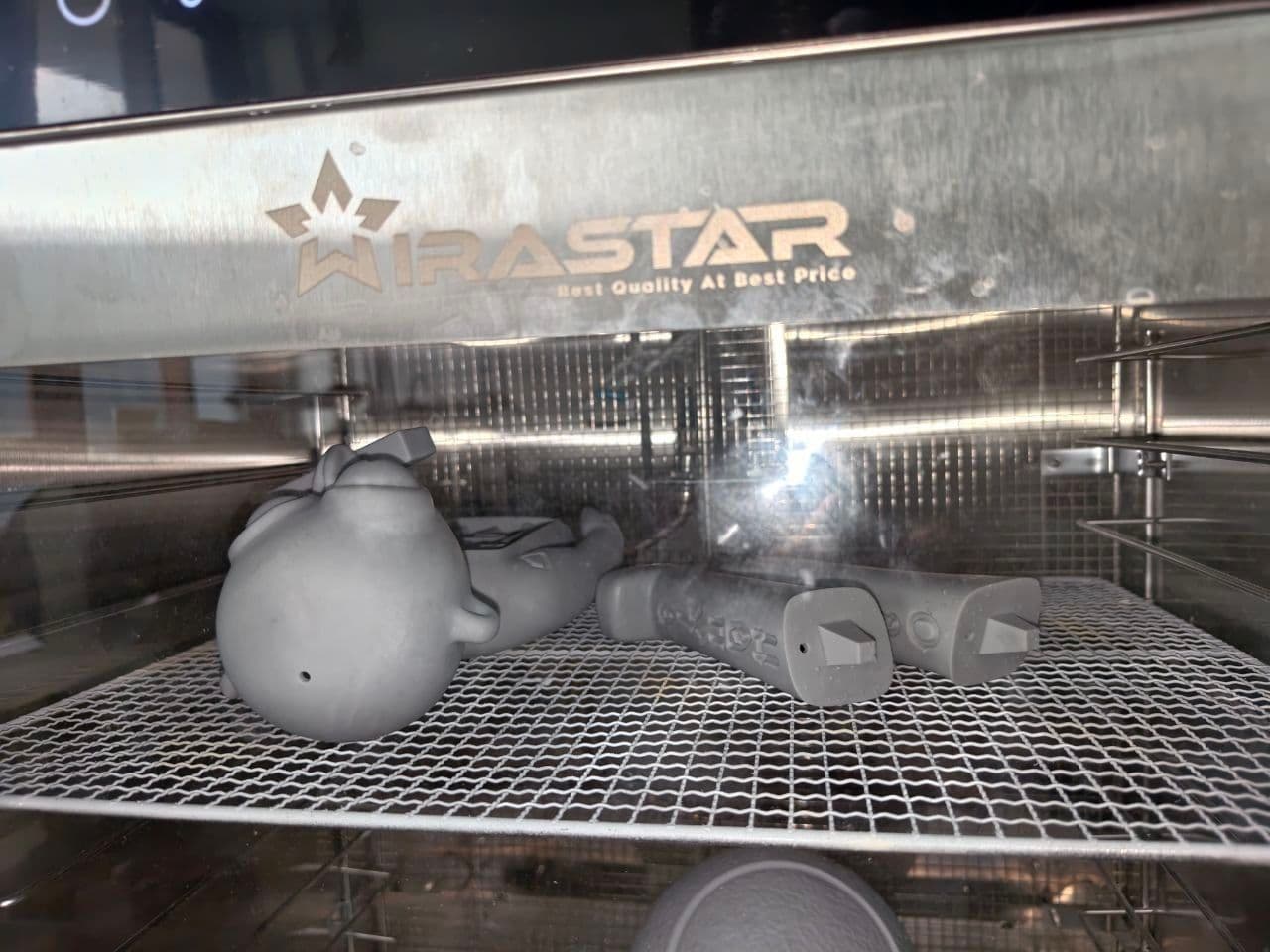 Sections of the Three Angels prototype curing on a wire rack inside a Wirastar dehydrator: the round gray resin head and glyph-marked leg pieces laid out under the branded steel panel.