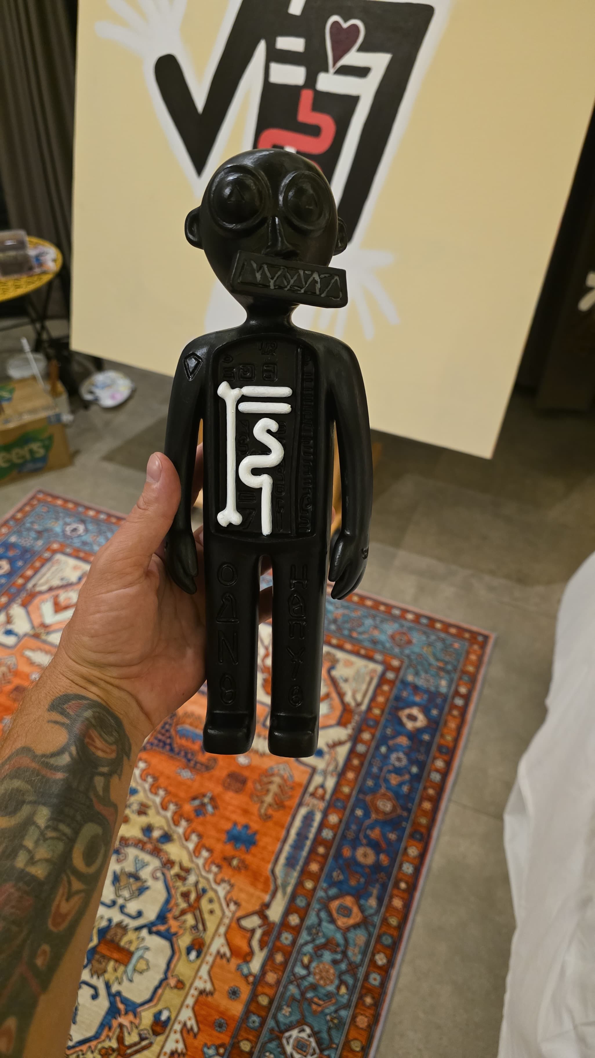 The Three Angels prototype mid hand-finishing, held up in the studio: body painted black with the bone and serpent chest markings picked out in white, glyph text running down the legs.