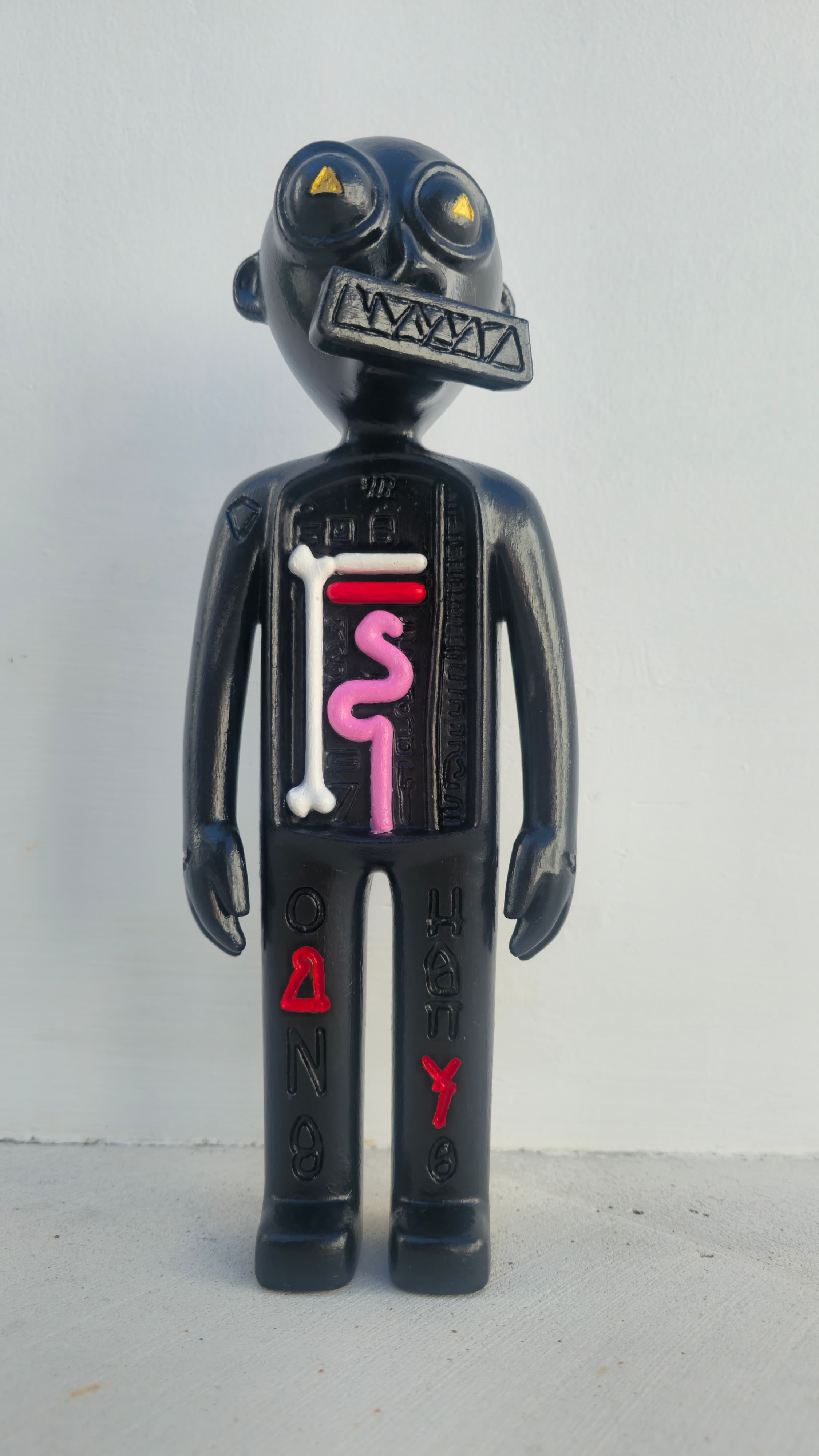 The finished Three Angels prototype standing in daylight against a white wall: body painted black, gold triangle pupils in the round eyes, the zigzag collar cut in silver-grey, the open torso carrying the white bone, red heart bar, and pink serpent, and the glyph rows down both legs picked out in white, red, pink, and yellow.