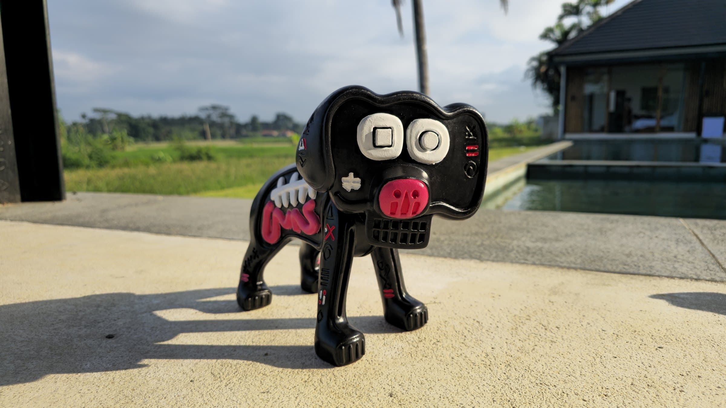 Marley by tijo (2026), a hand-painted 3D-printed resin talisman of the dog from the painting Boy & Dog V2: glossy black body, white-ringed square and circle eyes, a pink nose, a white tooth-grid, and white ribs with a pink intestinal coil painted down the flank. The soles are stamped 1/1 and signed tijo.