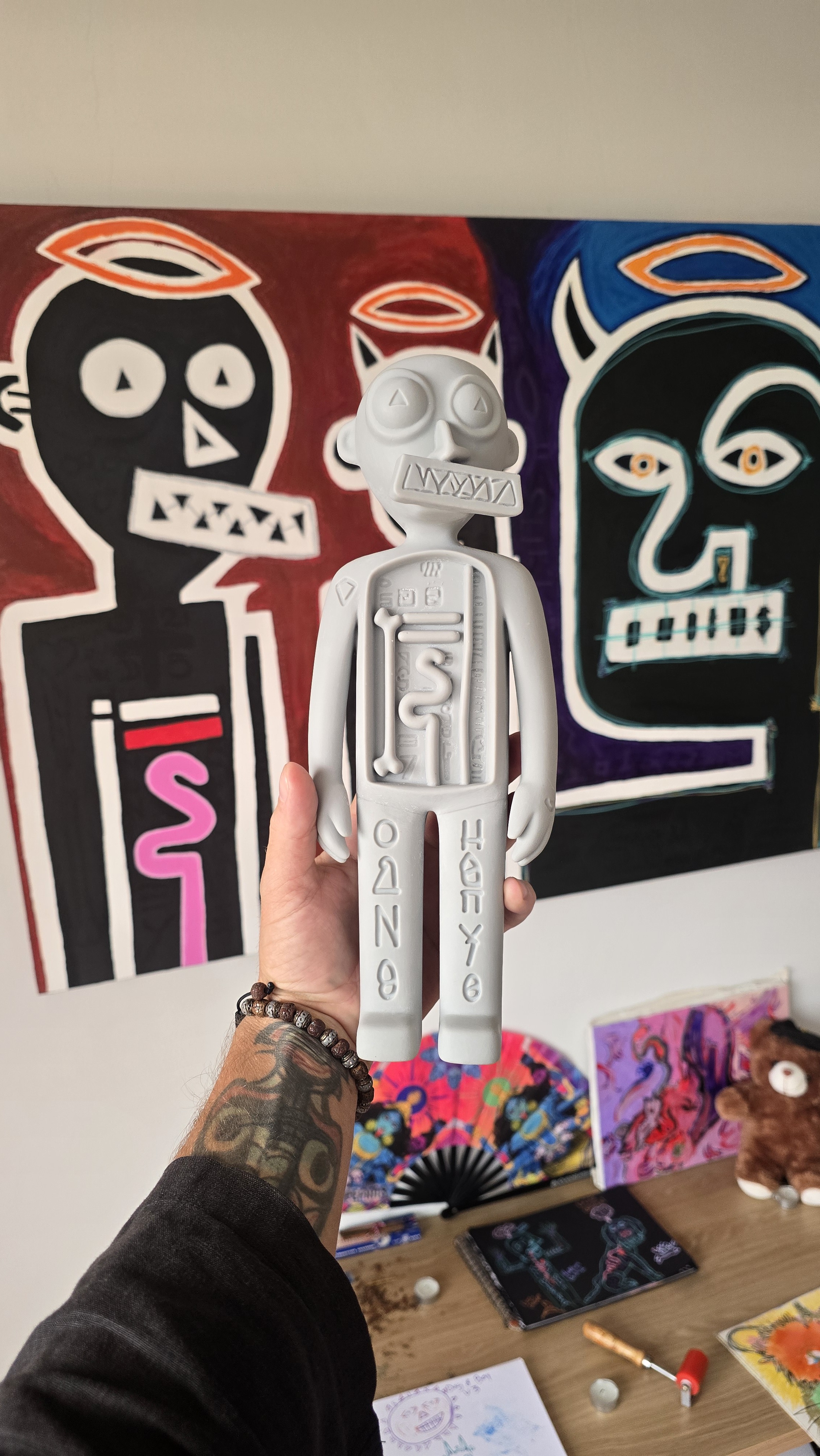 The Three Angels talisman held in the hand in front of its source painting — a twelve-inch painted guardian figure by tijo.